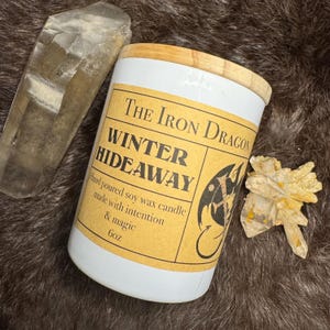 May include: A white candle with a wooden lid and a label that reads "The Iron Dragon WINTER HIDEAWAY." The label also states "Hand poured soy wax candle made with intention & magic 6oz." Two crystals are next to the candle.