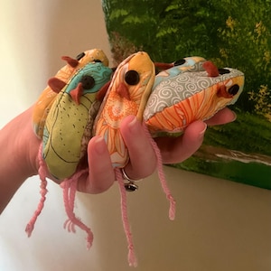 May include: A collection of handmade fabric creatures in shades of yellow, orange, and teal, with button eyes and pink yarn legs. The creatures are held in a hand, showcasing their unique patterns and textures.