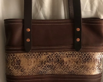 May include: A brown leather tote bag with a snakeskin pattern on the front. The bag has two brown leather straps and copper colored rivets.