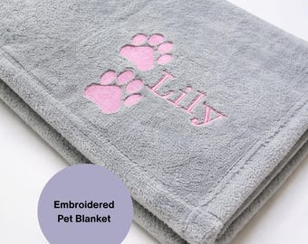 Pode incluir: Uma manta cinza dobrada para animais de estimação com pegadas rosa e o nome "Lily" bordado em rosa. A manta tem uma textura macia e felpuda. Um círculo roxo com o texto "Embroidered Pet Blanket" está no canto inferior esquerdo.