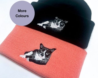 Puede incluir: Dos gorros de punto, uno negro y otro coral, están apilados. Cada gorro presenta un diseño de gato bordado en blanco y negro. Un círculo morado con el texto "More Colours" está en la esquina superior izquierda.
