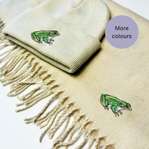 May include: A beige knitted beanie and scarf set with a green frog embroidered on each piece. The text "More colours" is on a purple circle in the top right corner of the image.