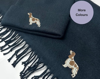 May include: A dark blue knitted beanie and scarf set with an embroidered brown and white dog design. The text "More Colours" is on a purple circle.