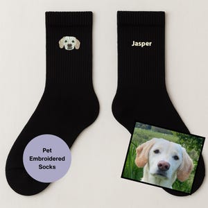 Op de afbeelding: Zwarte sokken met een geborduurd lichtgekleurd honden gezicht en de naam "Jasper". Een kleine foto van de hond is inbegrepen. De sokken zijn gelabeld "Pet Embroidered Socks".