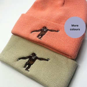 Puede incluir: Dos gorros de punto con un gráfico de mono bordado. El gorro de arriba es de color rosa coral y el gorro de abajo es beige. El texto "Más colores" está en un círculo morado en la esquina superior derecha de la imagen.