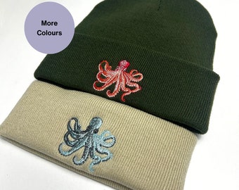 Pode incluir: Dois gorros de malha com desenhos de polvo bordados. O gorro de cima é verde escuro com um polvo vermelho. O gorro de baixo é bege com um polvo azul.