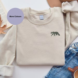 Puede incluir: Una sudadera beige con un pequeño gráfico de leopardo de las nieves en blanco y negro en el pecho izquierdo. La sudadera está doblada y colocada sobre una superficie blanca.