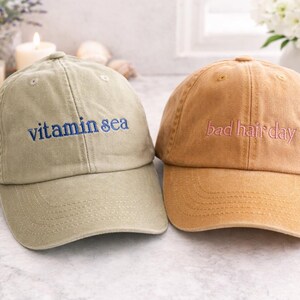 May include: Two baseball caps made of cotton fabric. The left cap is olive green with the text vitamin sea embroidered in blue. The right cap is tan with the text bad hair day embroidered in pink. Both caps feature a curved brim and eyelets.