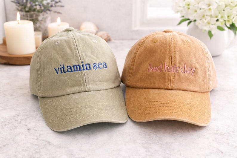 May include: Two baseball caps made of cotton fabric. The left cap is olive green with the text vitamin sea embroidered in blue. The right cap is tan with the text bad hair day embroidered in pink. Both caps feature a curved brim and eyelets.
