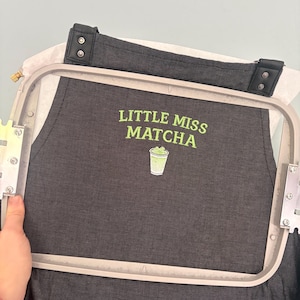 May include: Black apron with the text "LITTLE MISS MATCHA" in green, along with a green matcha drink graphic. The apron is held in an embroidery hoop, with a black strap and leather details.
