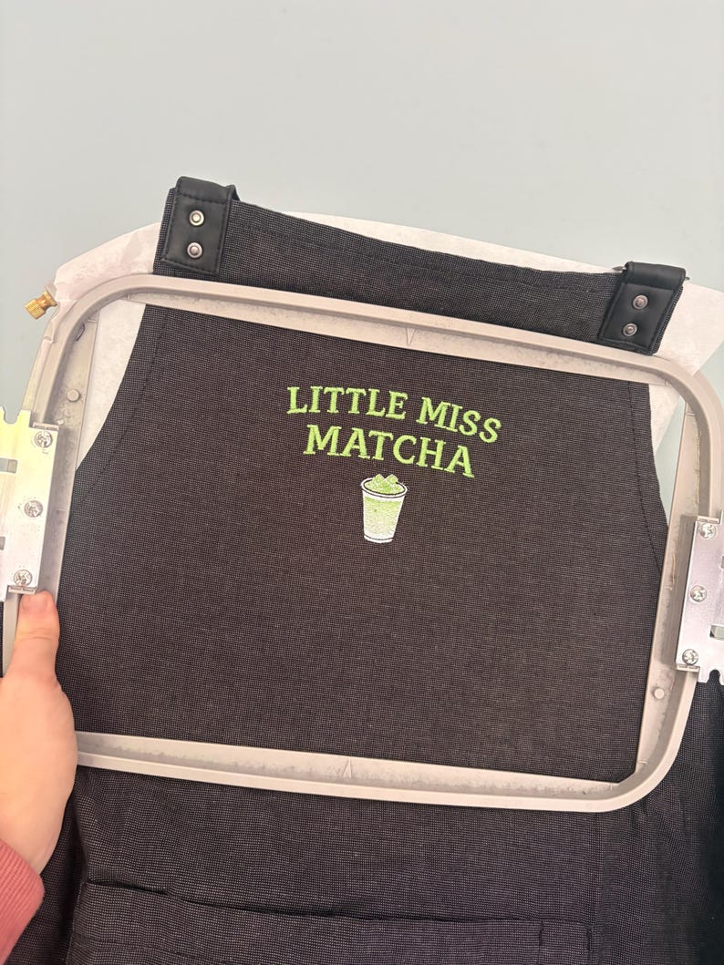 May include: Black apron with the text "LITTLE MISS MATCHA" in green, along with a green matcha drink graphic. The apron is held in an embroidery hoop, with a black strap and leather details.