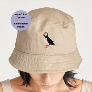 Puede incluir: Un sombrero de pescador beige con un diseño de frailecillo bordado. El sombrero tiene un ala ancha y una corona plana. Hay más opciones de color disponibles.