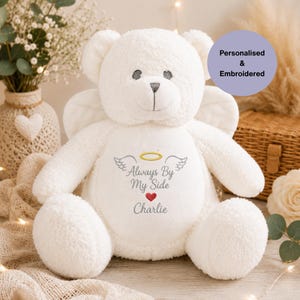 May include: A white plush teddy bear with angel wings and embroidered text on its chest that reads Always By My Side with a yellow halo, grey wings, a red heart, and the name Charlie. The bear has a soft, textured fabric finish.