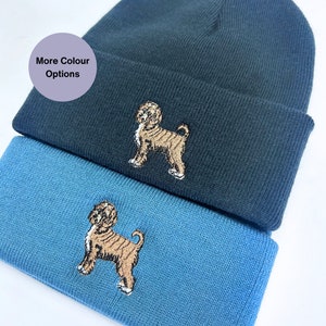 Könnte beinhalten: Zwei Strickmützen mit einem braunen Hund, der auf jeder Mütze gestickt ist. Die obere Mütze ist dunkelblau und die untere Mütze ist hellblau. Der Text "More Colour Options" befindet sich in einem violetten Kasten.