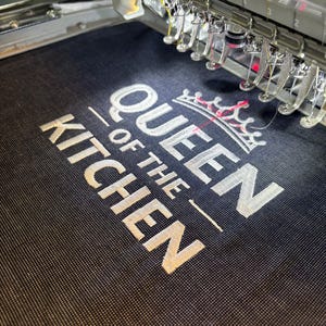 May include: Close-up of an embroidery machine stitching the words "QUEEN OF THE KITCHEN" in white thread onto a dark blue fabric. A crown design is above the text. The machine's needles and components are visible.