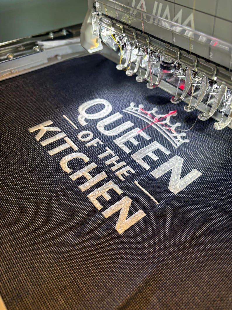 May include: Close-up of an embroidery machine stitching the words "QUEEN OF THE KITCHEN" in white thread onto a dark blue fabric. A crown design is above the text. The machine's needles and components are visible.