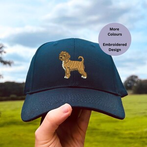 May include: A dark blue baseball cap with an embroidered design of a brown and white dog. The text "More Colours" and "Embroidered Design" is on a purple background.