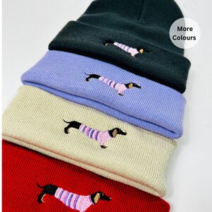 May include: A stack of four knitted beanies in different colours, each featuring an embroidered dachshund wearing a pink and white striped jumper. The beanie colours are black, lavender, beige, and red. The text "More Colours" is visible in the top right corner of the image.