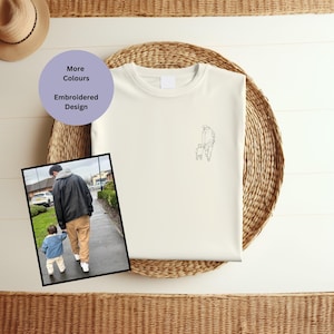 May include: A white t-shirt with an embroidered design of a father and child walking hand-in-hand. The design is in black outline. The t-shirt is on a woven surface. The text "More Colours" and "Embroidered Design" is on a purple circle.