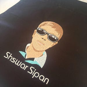 May include: Black apron with an embroidered portrait of a person wearing sunglasses. The portrait is in shades of brown, tan, and white. The name "Shswar Sipan" is embroidered in white below the portrait. The apron has a metal grommet at the top.