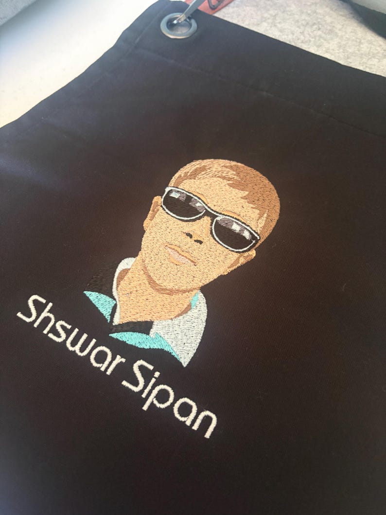 May include: Black apron with an embroidered portrait of a person wearing sunglasses. The portrait is in shades of brown, tan, and white. The name "Shswar Sipan" is embroidered in white below the portrait. The apron has a metal grommet at the top.