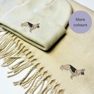 Puede incluir: Conjunto de gorro y bufanda de punto beige con un diseño de perro pastor alemán bordado. El conjunto está disponible en otros colores.