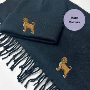Puede incluir: Un conjunto de gorro y bufanda de punto azul marino con un perro marrón bordado en cada pieza. La bufanda tiene flecos en el borde inferior. El texto "Más colores" está en un círculo morado.
