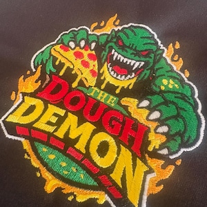 May include: Embroidered patch featuring a green monster with red eyes and sharp teeth, holding a slice of pizza. The text "THE DOUGH DEMON" is in red, yellow, and green, surrounded by flames. The patch is on a black background.