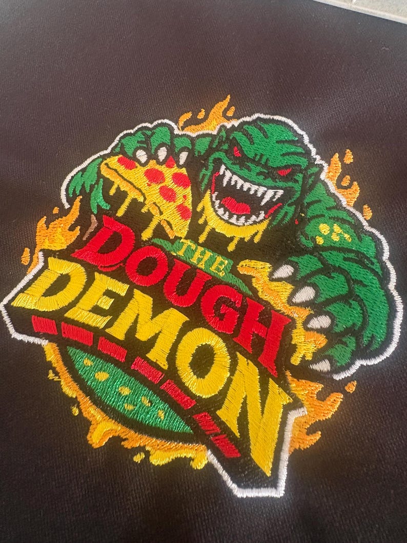 May include: Embroidered patch featuring a green monster with red eyes and sharp teeth, holding a slice of pizza. The text "THE DOUGH DEMON" is in red, yellow, and green, surrounded by flames. The patch is on a black background.
