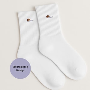 Puede incluir: Un par de calcetines blancos con diseño acanalado. Cada calcetín presenta un pequeño caracol bordado en marrón y gris. Los calcetines se muestran sobre un fondo claro. También se ve un círculo morado con el texto "Embroidered Design".