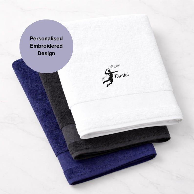 Puede incluir: Tres toallas bordadas personalizadas en blanco, negro y azul marino. La toalla blanca presenta un gr&aacute;fico de jugador de b&aacute;dminton negro y el nombre "Daniel". Las toallas est&aacute;n dobladas y apiladas, mostrando un dise&ntilde;o limpio y moderno.
