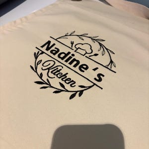 May include: Cream-colored apron with the text "Nadine's Kitchen" in black script, encircled by a wreath design. The apron features a chef's hat and utensils graphic. It has a brown leather strap and a cream-colored neck strap.