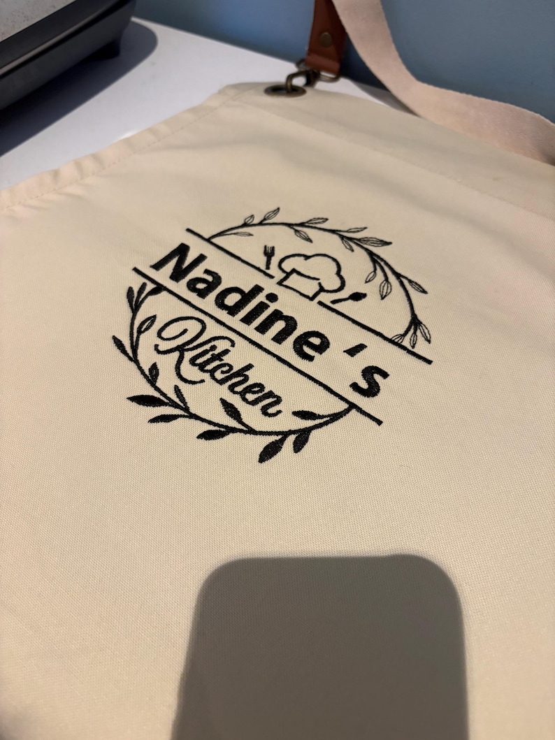 May include: Cream-colored apron with the text "Nadine's Kitchen" in black script, encircled by a wreath design. The apron features a chef's hat and utensils graphic. It has a brown leather strap and a cream-colored neck strap.
