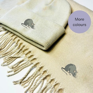 Peut inclure: Un bonnet en tricot beige et une écharpe assortie avec un motif de rat brodé gris. Le texte "Plus de couleurs" est sur un cercle violet.