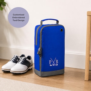 Puede incluir: Una bolsa de zapatos de golf azul real con ribete gris y un asa, junto a un par de zapatos de golf blancos y azul marino. La bolsa tiene una cremallera y las iniciales "PXB" con palos de golf cruzados. El texto en la imagen dice "Customised Embroidered Text/Design."