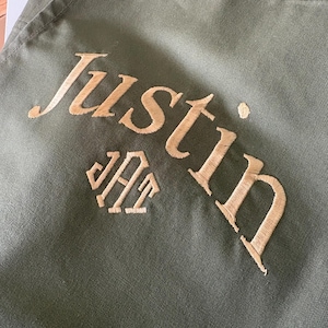 May include: Olive green fabric with the name "Justin" embroidered in a gold-colored thread. Below the name is a monogram in the same gold thread. The fabric has a slightly textured appearance.