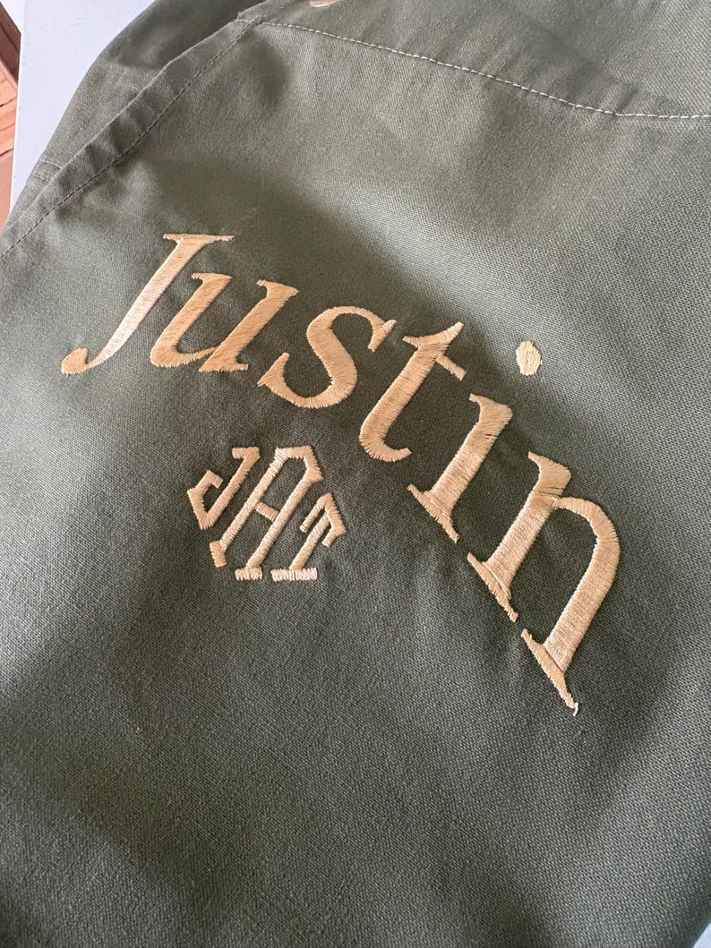 May include: Olive green fabric with the name "Justin" embroidered in a gold-colored thread. Below the name is a monogram in the same gold thread. The fabric has a slightly textured appearance.