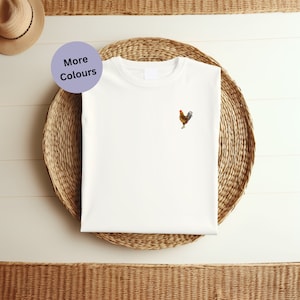 May include: White t-shirt with a small embroidered rooster design. The rooster is brown and yellow with a red comb.