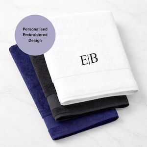 Puede incluir: Tres toallas personalizadas con dise&ntilde;o bordado en blanco, negro y azul marino. La toalla blanca tiene las iniciales "EB" bordadas. Las toallas est&aacute;n dobladas y apiladas, con la toalla blanca encima.