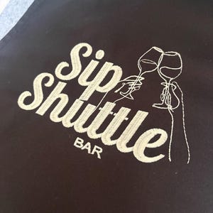May include: Black apron with the embroidered text "Sip Shuttle Bar" in white script. The design includes line art of two wine glasses clinking, suggesting a bar or wine-related theme.
