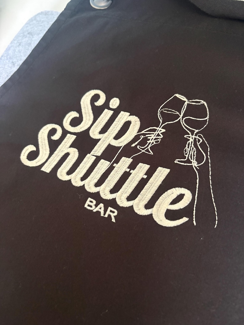 May include: Black apron with the embroidered text "Sip Shuttle Bar" in white script. The design includes line art of two wine glasses clinking, suggesting a bar or wine-related theme.