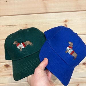 Puede incluir: Dos gorras de b&eacute;isbol, una de color verde oscuro y otra azul real, cada una con un perro salchicha marr&oacute;n bordado que lleva un su&eacute;ter blanco y rojo. Las gorras tienen una corona estructurada, visera curva y ojales de ventilaci&oacute;n.