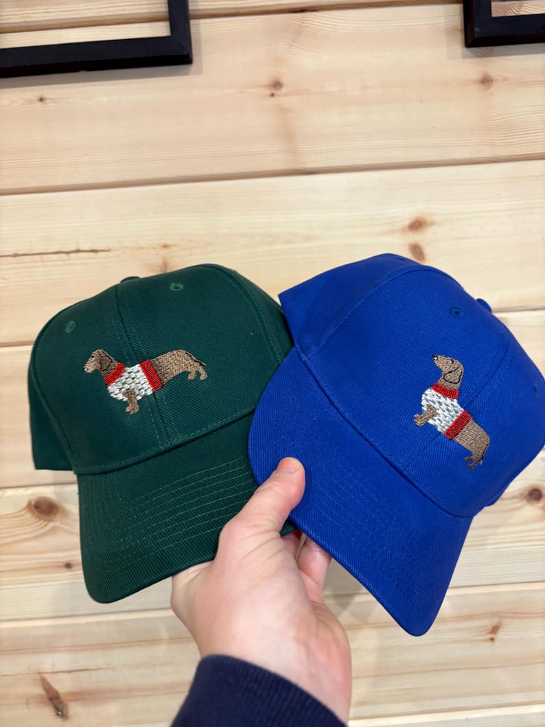 Puede incluir: Dos gorras de b&eacute;isbol, una de color verde oscuro y otra azul real, cada una con un perro salchicha marr&oacute;n bordado que lleva un su&eacute;ter blanco y rojo. Las gorras tienen una corona estructurada, visera curva y ojales de ventilaci&oacute;n.