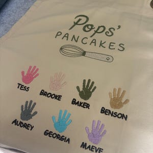 May include: Beige apron with the text "Pops' Pancakes" and a whisk illustration. Features embroidered handprints in various colors with names: Tess, Brooke, Baker, Benson, Audrey, Georgia, and Maeve. Includes brass grommets.