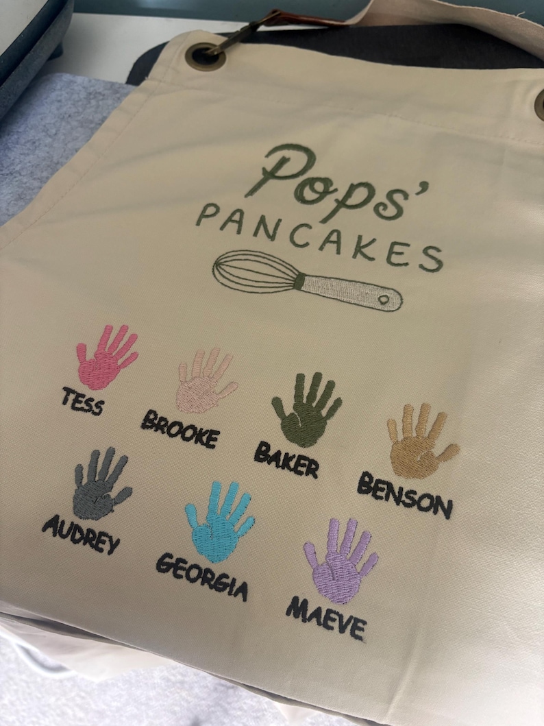 May include: Beige apron with the text "Pops' Pancakes" and a whisk illustration. Features embroidered handprints in various colors with names: Tess, Brooke, Baker, Benson, Audrey, Georgia, and Maeve. Includes brass grommets.