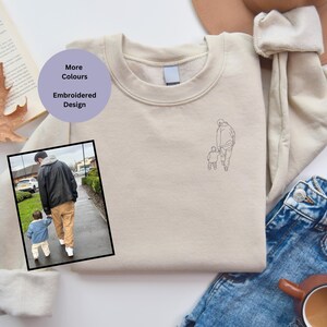 Puede incluir: Una sudadera beige con un dibujo lineal de un padre y un hijo caminando de la mano bordado en la parte delantera. El texto "More Colours" y "Embroidered Design" está escrito en un círculo morado en la parte superior izquierda de la imagen.