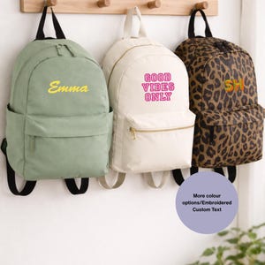 May include: Three backpacks hanging on wooden pegs. One is sage green with "Emma" in yellow script, another is off-white with "GOOD VIBES ONLY" in pink, and the third has a leopard print with "SH" in rainbow colors. All have front pockets.
