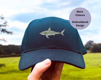 May include: A navy blue baseball cap with an embroidered gray shark design. The text "More Colours" and "Embroidered Design" is on a purple circle.