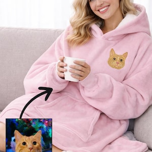 May include: A pale pink hooded blanket featuring an embroidered cat face. The blanket has a large pocket and a white fleece lining in the hood. The person is holding a white mug.