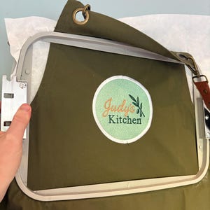 May include: Olive green apron with a white and orange embroidered logo that reads "Judy's Kitchen" with olive branch accents. The apron is in an embroidery hoop, ready for stitching. Features a brown leather strap and metal grommets.